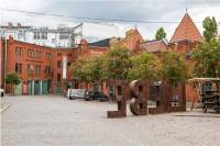 A large terracotta bricked building is visible across a courtyard. On the ground floor, the building has green doors. There are six trees in bloom in the courtyard. Two cars are parked underneath them. The word 