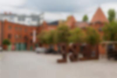 A large terracotta bricked building is visible across a courtyard. On the ground floor, the building has green doors. There are six trees in bloom in the courtyard. Two cars are parked underneath them. The word 