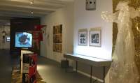 Exhibition space with several installations. At the back, there is an image being projected on a wall, and a large doll dressed as a jester. There are photographs on the side wall and a table emitting some light. A plastic shape, resembling a dress, hangs next to it. At the centre of the space, another table has a propped up brochure.