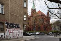 Empty street. Two buildings are visible, a brown building and a terracotta one, with a green tower. There are several cars parked in front of the second building and some bare trees along the street. The sky is cloudy.