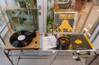 On top of a glass table there is a vinyl player, with the needle on on top of the vinyl disk. To the right, there is an open record jacket with headphones on top. Behind it, another album cover of the same record has been propped up.
