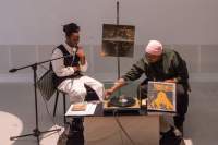 Two people sit behind a low table. On the table, there is a vinyl player, an album propped up and some paper. There is a microphone in front of the person on the left. The person on the right adjusts the needle of the record player. Behind them, in the middle of the two, there is a pole where a square photograph has been attached.