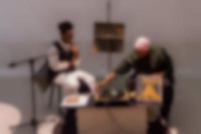 Two people sit behind a low table. On the table, there is a vinyl player, an album propped up and some paper. There is a microphone in front of the person on the left. The person on the right adjusts the needle of the record player. Behind them, in the middle of the two, there is a pole where a square photograph has been attached.