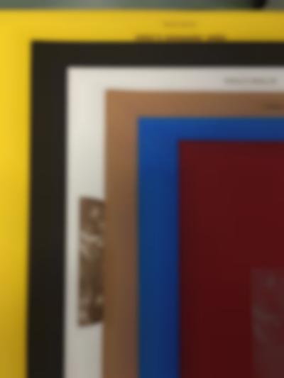 Several print materials have been stacked on top of each other, with the edges visible. Each sheet has a different colour; from bottom to top: yellow, black, white, light brown, blue and burgundy.