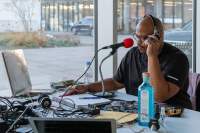 A person sits at a table, in front of a large red microphone. They are wearing headphones. On the table, there are several wires, an open notebook and a pen, a blue bottle, an Evian bottle and several other objects. Behind them, a courtyard and two cars are visible through the large windows.