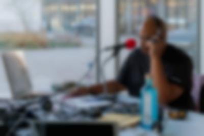 A person sits at a table, in front of a large red microphone. They are wearing headphones. On the table, there are several wires, an open notebook and a pen, a blue bottle, an Evian bottle and several other objects. Behind them, a courtyard and two cars are visible through the large windows.