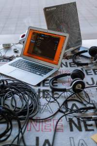 On top of a table, there is a laptop, two headphones, several wires and a vinyl cover. Under the items, there is a table cloth with the words 