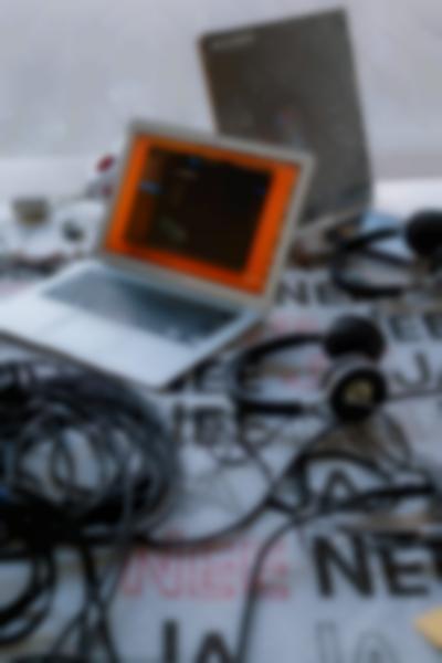 On top of a table, there is a laptop, two headphones, several wires and a vinyl cover. Under the items, there is a table cloth with the words 