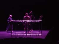 On stage, three people appear behind a metal structure. Purple light lights the space.