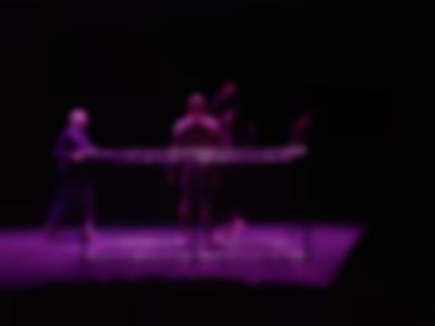 On stage, three people appear behind a metal structure. Purple light lights the space.