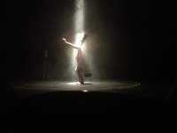 A person wearing a tall hat and a cape is at the center of the stage, with one of their arms raised. A spotlight shines on top of them.