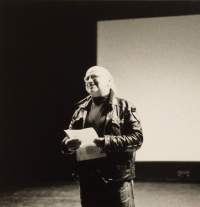 Black-and-white portrait of a person on stage. They are wearing a leather jacket and holding a sheet of paper. The person is looking up and smiling.