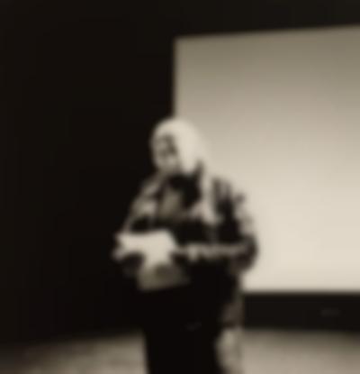 Black-and-white portrait of a person on stage. They are wearing a leather jacket and holding a sheet of paper. The person is looking up and smiling.