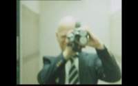 A person wearing a suit and tie holds a camera at their eye. The image is slightly blurry.