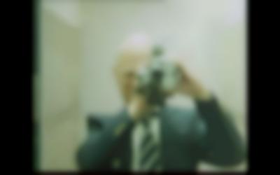 A person wearing a suit and tie holds a camera at their eye. The image is slightly blurry.