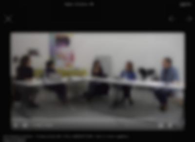Video player on top of a black background. There is an X on the top left corner of the page and two arrows in opposite directions on the right. Inside the video player, the visible still shows five people sitting around a L shape table. They each have a microphone in front of them.
