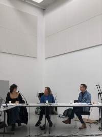 Three people sit behind a table. They each have a microphone in front of them. The two people on the right look in the direction of the person on the left, who is looking back at them.