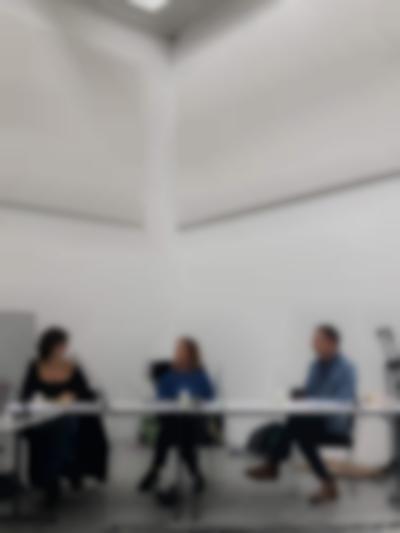 Three people sit behind a table. They each have a microphone in front of them. The two people on the right look in the direction of the person on the left, who is looking back at them.