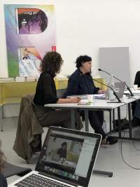 Two people sit at a table with microphones in front of them. There is an open laptop in between them. In the foreground, an open laptop is visible on someone's lap. The image on the laptop shows a close-up image of one of the people sitting at the table.