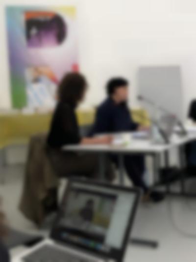 Two people sit at a table with microphones in front of them. There is an open laptop in between them. In the foreground, an open laptop is visible on someone's lap. The image on the laptop shows a close-up image of one of the people sitting at the table.