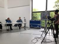 On the left, two people are sitting behind a table with microphones in front of them. On the right, a person stands behind two tripods with cameras. To their left, a screen is mounted on a stand. There are large windows at the back of the room and a blue equipment box.