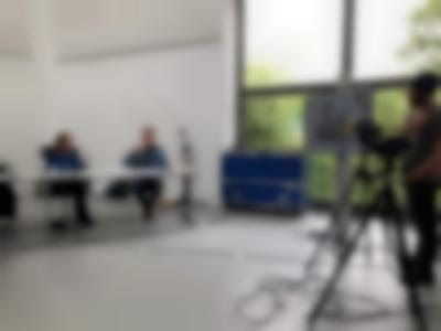 On the left, two people are sitting behind a table with microphones in front of them. On the right, a person stands behind two tripods with cameras. To their left, a screen is mounted on a stand. There are large windows at the back of the room and a blue equipment box.