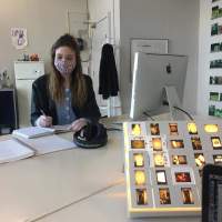 A person sits at a table, in front of an Apple desktop computer. They are writing on a notebook. Next to the notebook, there is an open spiral bound book and a pair of headphones. At the forefront, part of an open laptop computer is visible, as well as a lit up box with several slide photographs set on top. At the back of the room, behind the person writing, there is a fridge and a jacket in a coat stand.