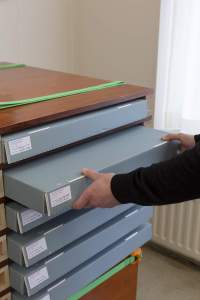 In a wooden shelf cabinet, there are 6 grey storage boxes with white stickers on the left side. A person, whose hands and forearms are visible, is pulling out the second box from the top.