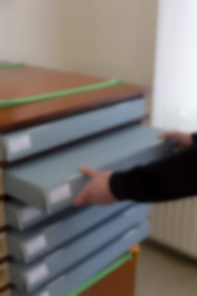 In a wooden shelf cabinet, there are 6 grey storage boxes with white stickers on the left side. A person, whose hands and forearms are visible, is pulling out the second box from the top.