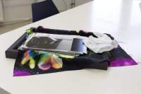 A piece of black and purple fabric with a flower lies extended on top of a black frame, on top of a white table. On top of the fabric, there is a stack of photographs and a pair of white gloves. A chair is partially visible at the back, next to the table.