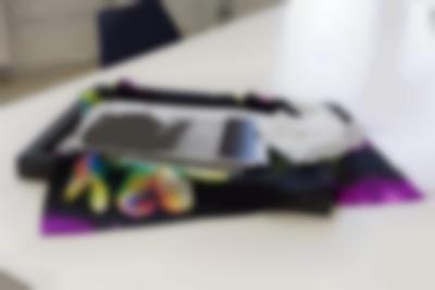 A piece of black and purple fabric with a flower lies extended on top of a black frame, on top of a white table. On top of the fabric, there is a stack of photographs and a pair of white gloves. A chair is partially visible at the back, next to the table.