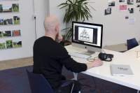 A person sits in front of an Apple desktop computer. They have their left hand on their chin and the right hand on the computer mouse. On the table, there is a pair of headphones and a closed spiral bound book. They are looking at a photograph on the desktop screen, which shows four frames hanging on a wall: three rectangular ones and, above them, a triangular one. At the back, there are four rows of photograph shelfs with photographs of green, natural settings; a tall plant and several photographs attached to the wall.