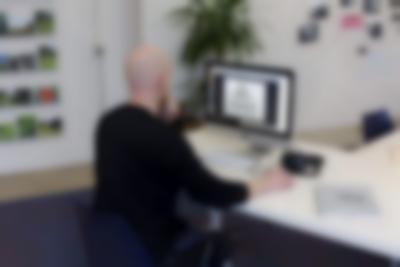 A person sits in front of an Apple desktop computer. They have their left hand on their chin and the right hand on the computer mouse. On the table, there is a pair of headphones and a closed spiral bound book. They are looking at a photograph on the desktop screen, which shows four frames hanging on a wall: three rectangular ones and, above them, a triangular one. At the back, there are four rows of photograph shelfs with photographs of green, natural settings; a tall plant and several photographs attached to the wall.