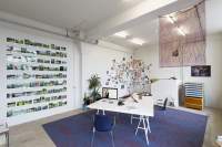Indoor space with white walls and a large purple carpet at the centre. On top of the carpet, there is a L shape white table, with two chairs. On top of the table there is a desktop computer, headphones, a book and other objects. Hanging on the wall, to the left, are nine rows of photograph shelfs, filled with photographs. To the right, there is a tall plant and next to it a large word gratified in brown ink, across the corner of the room. Hanging on top of the graffiti, there is an arrangement of photographs. A white block and a wooden cabinet are visible at the back of the room. A photograph hangs above the cabinet. Hanging from the ceiling, in the middle of the room, there is a piece of see-through purple fabric with words painted in gold.