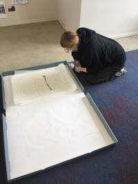 A person sits on their heels. In front of them, there is an open storage box with a stack of A3 paper. The person is leaning towards this paper, looking down at it. They wear all black, glasses and a face mask.