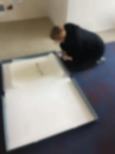 A person sits on their heels. In front of them, there is an open storage box with a stack of A3 paper. The person is leaning towards this paper, looking down at it. They wear all black, glasses and a face mask.