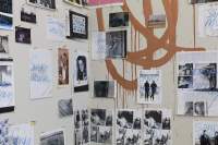 A scribble has been graffitied on a wall in light brown ink. Several photographs and printed pages have been nailed to the wall, some of them partially covering the scribble. Most of these images have 