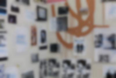 A scribble has been graffitied on a wall in light brown ink. Several photographs and printed pages have been nailed to the wall, some of them partially covering the scribble. Most of these images have 