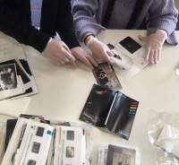 On top of a white surface, there are several photographs and photo envelopes. The hands and arms of two people are visible. They are wearing white gloves and handling the pictures. One of them is putting a stack of photographs inside an envelope, and another touches one of the photographs on the table.