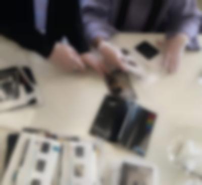 On top of a white surface, there are several photographs and photo envelopes. The hands and arms of two people are visible. They are wearing white gloves and handling the pictures. One of them is putting a stack of photographs inside an envelope, and another touches one of the photographs on the table.
