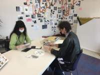 Two people sit across from each other at a long white table. They are both wearing white gloves and holding photographs, which they are looking at. There are photo envelopes on the table, a see-through plastic bag and a round white plate with two stars and a word written in black. At the back of the room, there are several photographs on the wall. Behind them, there is a large hand-painted word, which is not fully visible.
