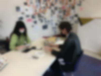 Two people sit across from each other at a long white table. They are both wearing white gloves and holding photographs, which they are looking at. There are photo envelopes on the table, a see-through plastic bag and a round white plate with two stars and a word written in black. At the back of the room, there are several photographs on the wall. Behind them, there is a large hand-painted word, which is not fully visible.