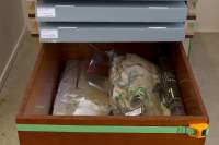 Wooden box with two plastic bags with paper and fabric inside. Above the box. There are two grey storage boxes with white stickers on the left side.