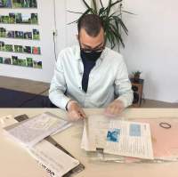 A person sits at a table. In front of them, there are two small stacks of printed materials. They are wearing white gloves and handling one of the sheets of paper. Behind the person, there is a tall plant. To the left, there are 5 rows of photograph shelfs with photographs of green, natural settings.