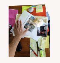 On top of a table, there are several sticky notes, a photograph, a shell, a rock, pens and highlighters, and a sheet of paper with a photograph of two people and some words in black text. A hand is resting on top of this paper. There is a white digital frame around the photograph.