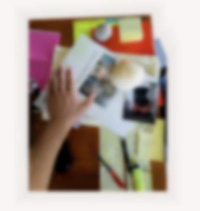 On top of a table, there are several sticky notes, a photograph, a shell, a rock, pens and highlighters, and a sheet of paper with a photograph of two people and some words in black text. A hand is resting on top of this paper. There is a white digital frame around the photograph.
