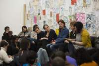 Eight people sit in line. The person on the right is holding paper and looking down at it. The other people look in their direction. Behind them, there is a wall with drawings and hand-drawn posters. A group of people sits on the floor, facing the eight people who sit on chairs.