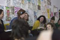 A few people sit in line. The three people in the middle are looking at each other and laughing. Behind them, there is a wall with drawings and hand-drawn posters. Two other people, whose heads are visible, are sitting on the floor in front of them.