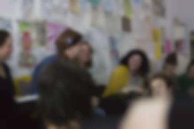 A few people sit in line. The three people in the middle are looking at each other and laughing. Behind them, there is a wall with drawings and hand-drawn posters. Two other people, whose heads are visible, are sitting on the floor in front of them.