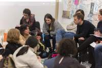 A few people sit in line. Two of them are leaning forward, supporting their elbows on their knees. They lean towards the people who are sitting on the floor, facing them. One of the people on the floor is gesturing with their hands. The other people look towards them.